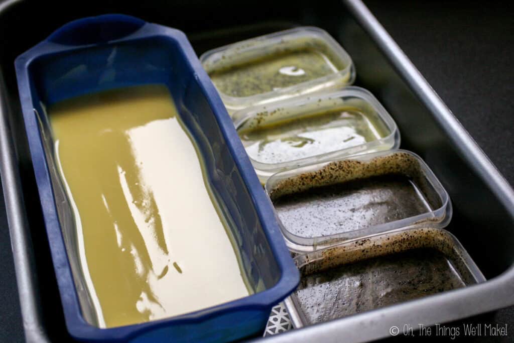 A soap mixture in a silicone loaf pan, and soap mixtures with coffee grains and poppy seeds in smaller plastic containers