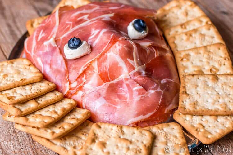 Prosciutto face with mozzarella cheese ball eyes and blueberry pupils on a platter surrounded by crackers
