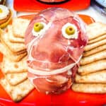 Creepy Halloween dish shaped as a head made out of Prosciutto ham with cream cheese and green olive eyes, surrounded by square crackers.