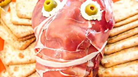 Creepy Halloween dish shaped as a head made out of Prosciutto ham with cream cheese and green olive eyes, surrounded by square crackers.