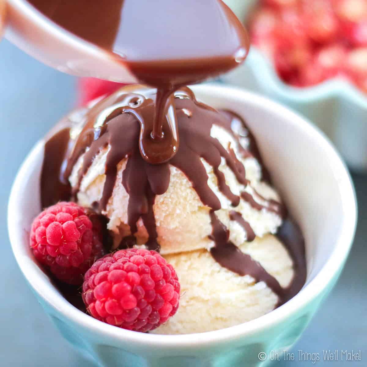 Close-up of homemade chocolate shell sauce being pouring over some vanilla ice cream with raspberries, inside a light blue bowl.