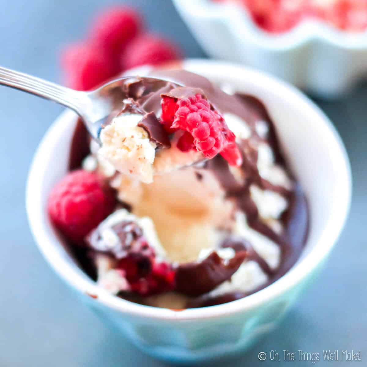 Closeup of a spoonful of frozen kefir ice cream with chocolate magic shell and part of a raspberry, over a bowl of frozen kefir covered with chocolate magic shell.