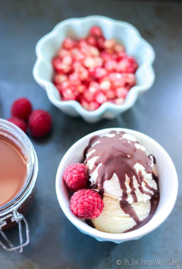 Overhead view of a bowl of ice cream covered with a hard chocolate topping and garnished with raspberries. Behind it, there is a bowl of pomegranate seeds. To its left, there is a jar of liquid chocolate.