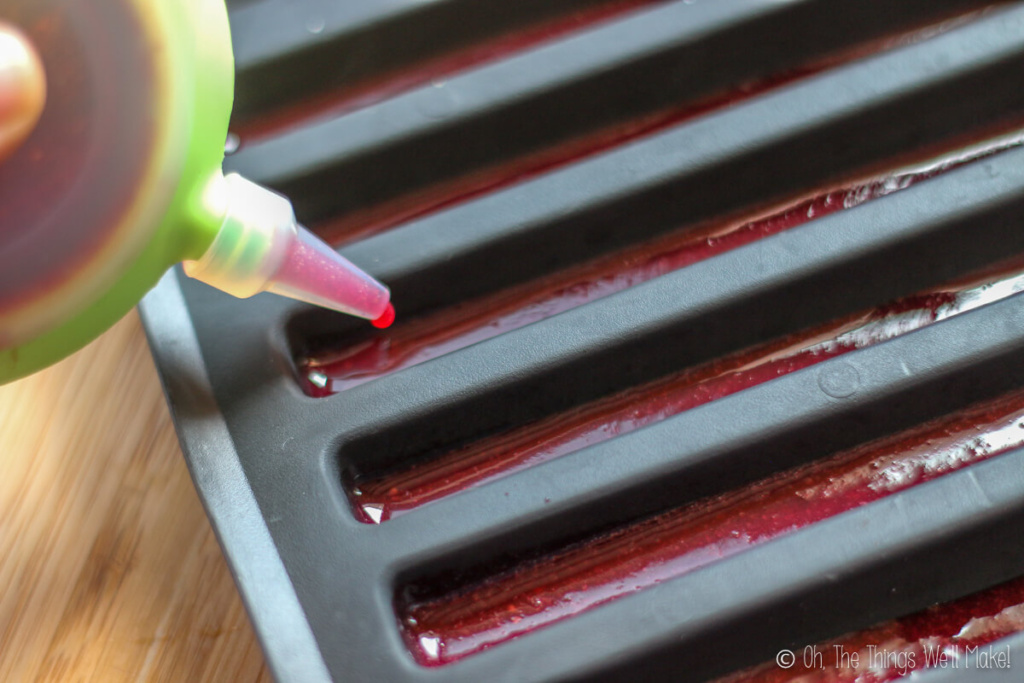 Close up of a silicone bottle pouring red gelatin into a ice cube tray mold.