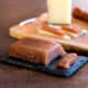 A block of homemade sweet quince paste (dulce de membrillo) on a black rectangular platter, in front of a cutting board with manchego cheese and chorizo sausage.