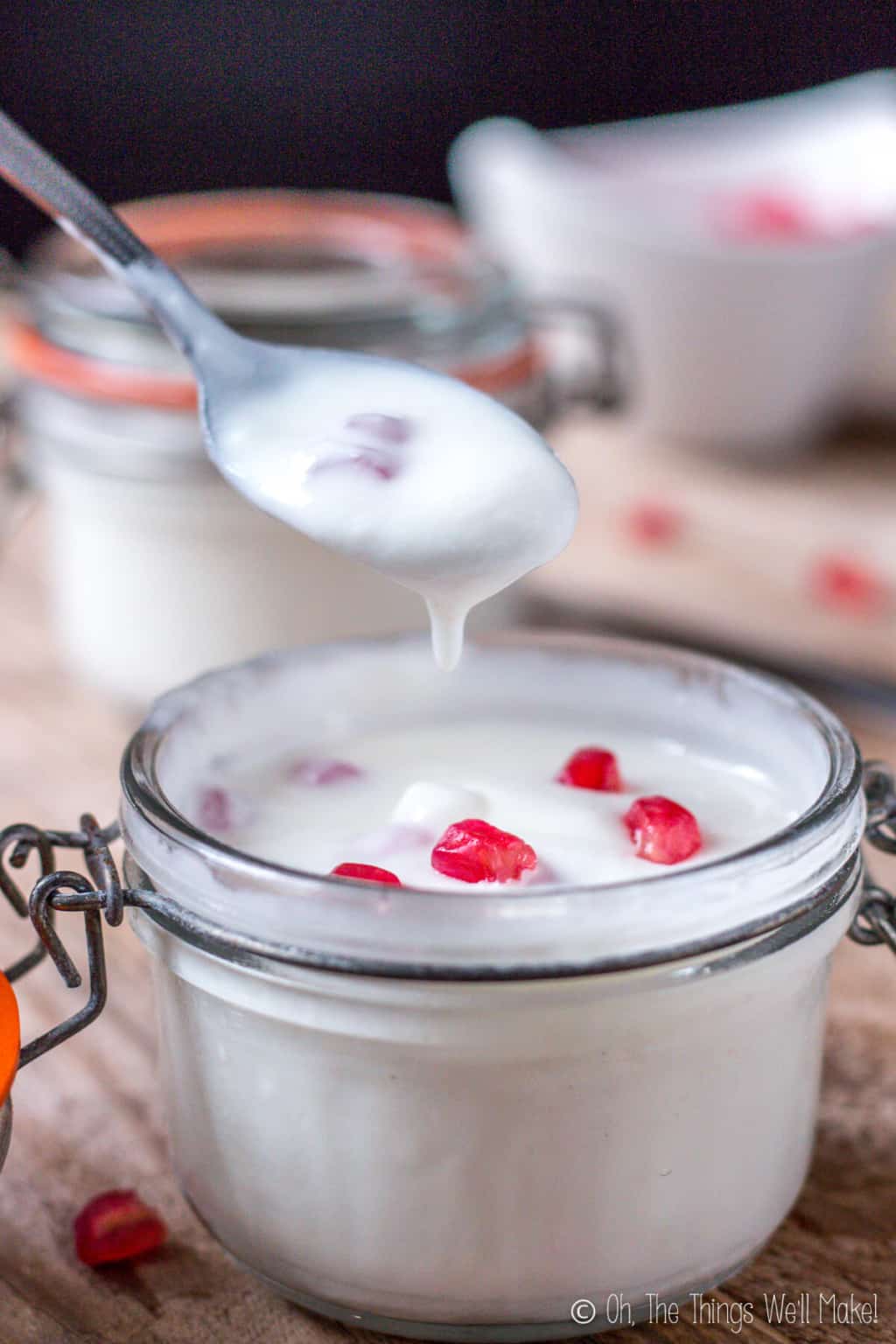 Yogurt on a spoon above a glass jar of yogurt with pomegranate seeds. There is some yogurt slightly dripping back down off the spoon showing the texture. 