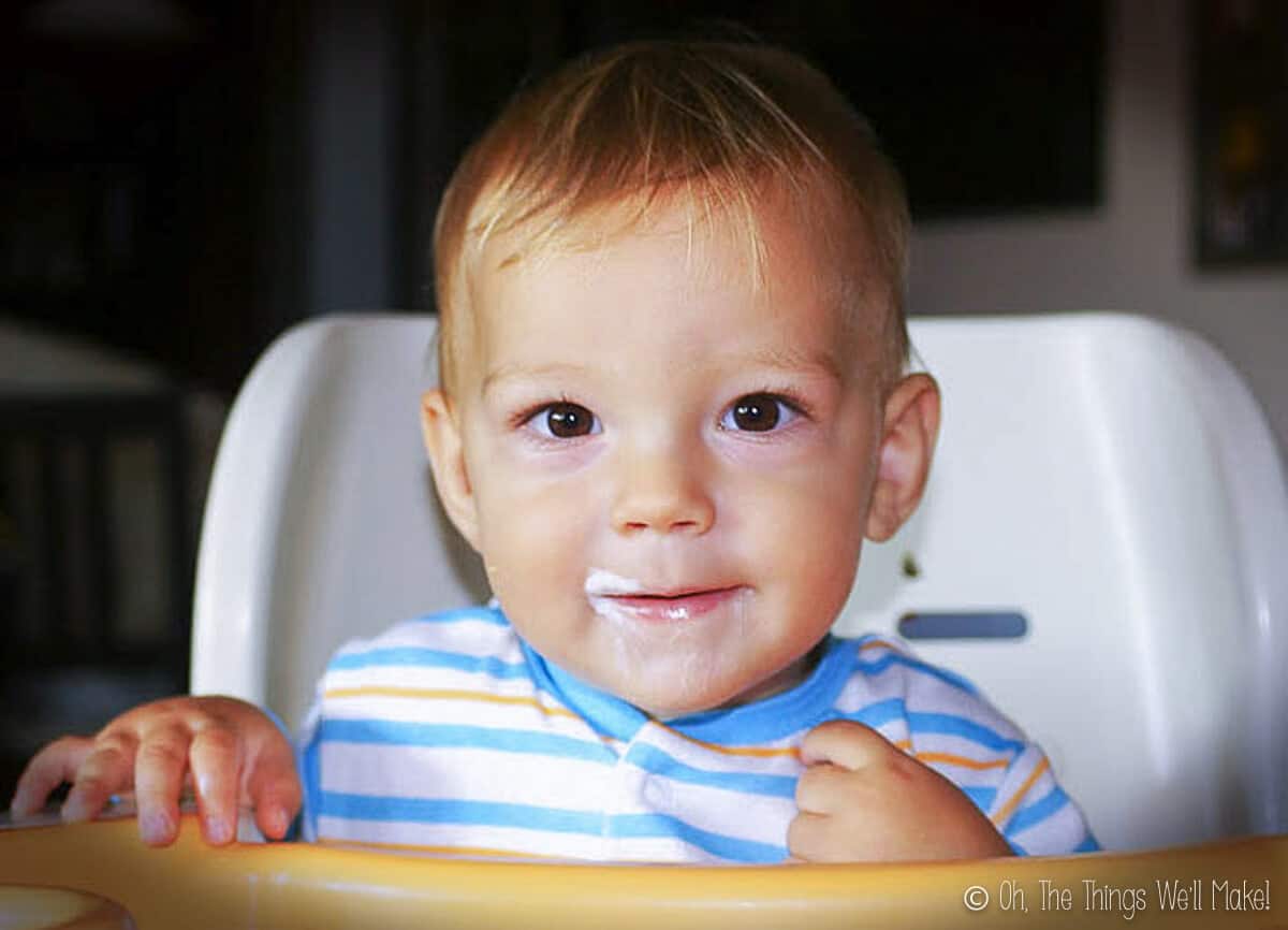 A baby boy sitting in a high chair with yogurt on his face. 