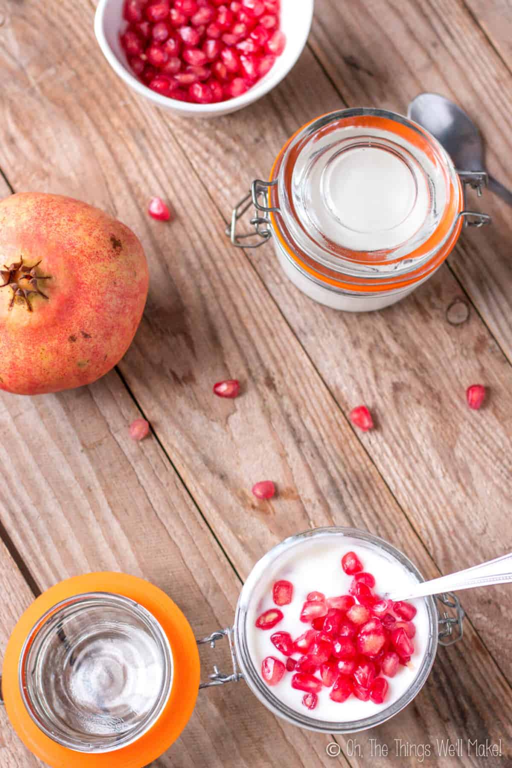 Overhead view of two jars of homemade yogurt on a wood surface. Bottom jar is open and has a spoon inside it with fresh pomegranate topping it. Top jar is closed and is beside a pomegranate fruit and a bowl full of pomegranate seeds.