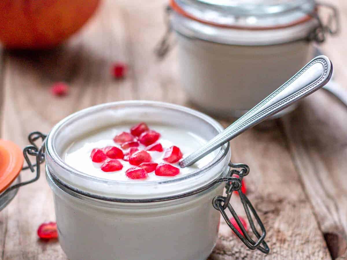 Close-up of two jars full of yogurt. One jar open showing yogurt garnished with pomegranate seeds and a spoon inside it.