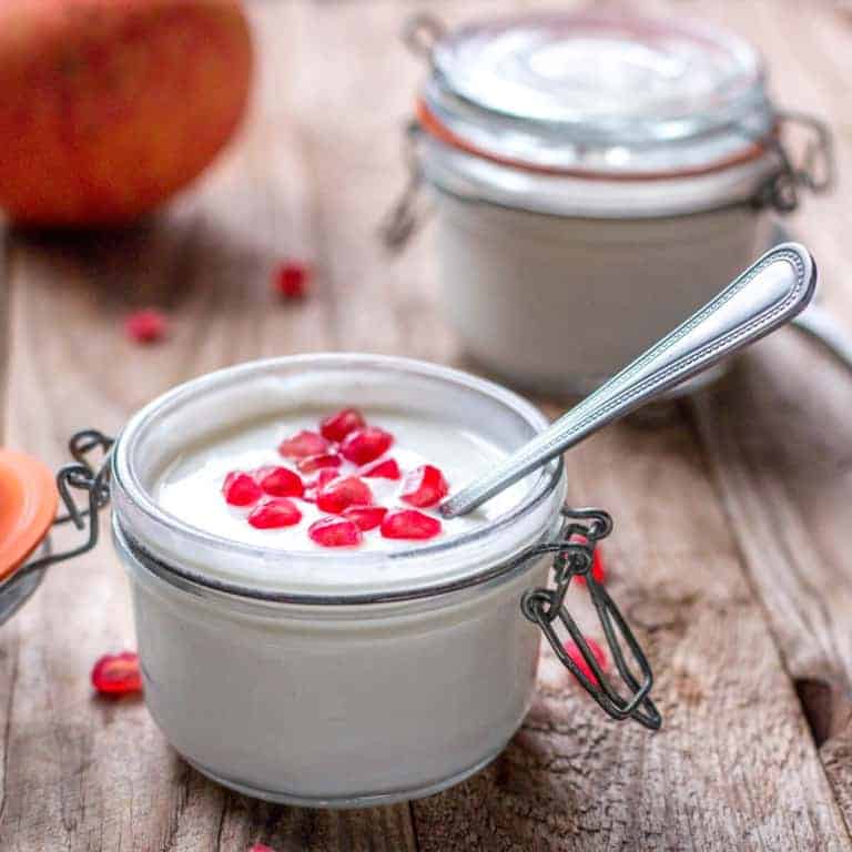 Close-up of two jars full of yogurt. One jar open showing yogurt garnished with pomegranate seeds and a spoon inside it.