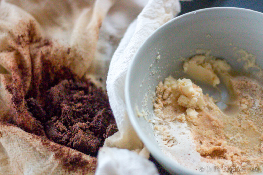 Close up of dark brown foamy milk solids on a cheesecloth and a white bowl on the right, filled with crunchy milk solids from making ghee.