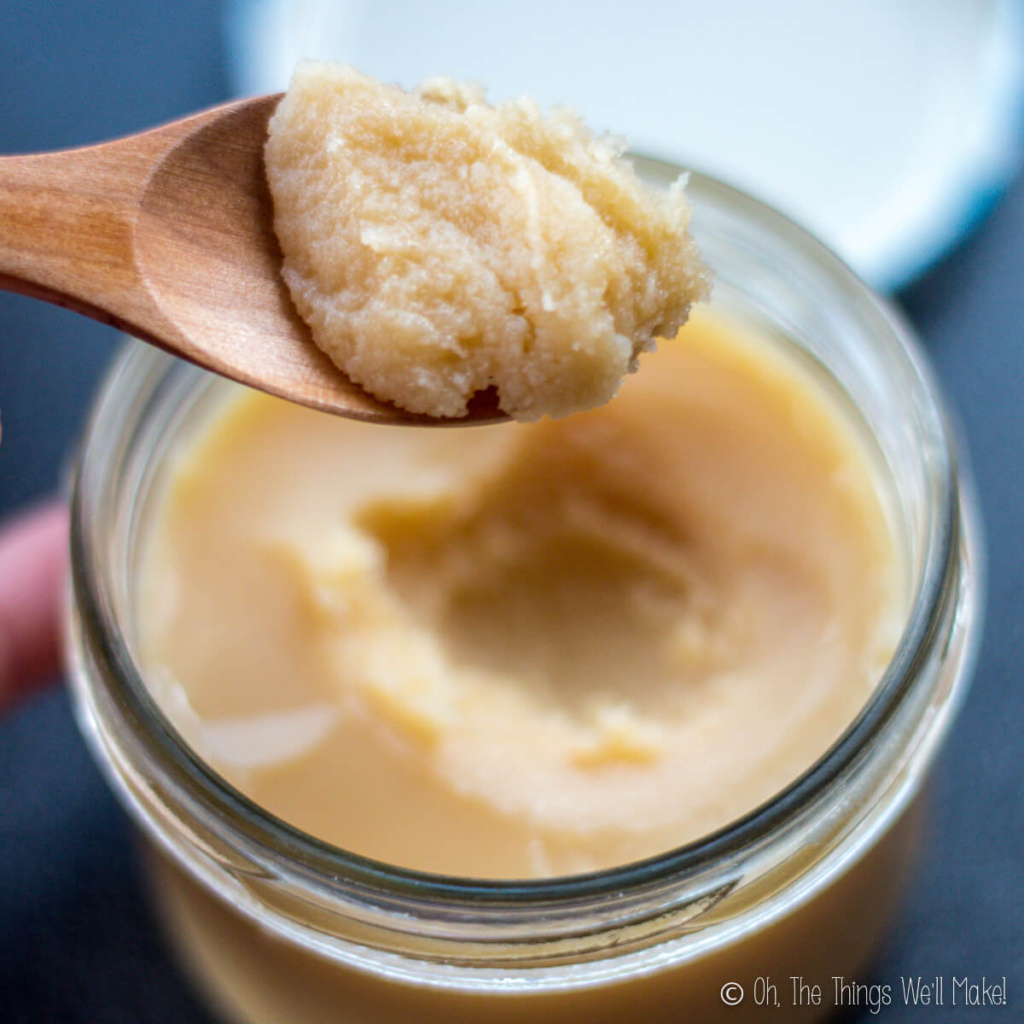Jar of ghee that is solid after being stored in the fridge, with a spoon of the solid ghee held up over the jar to show a closeup of the texture