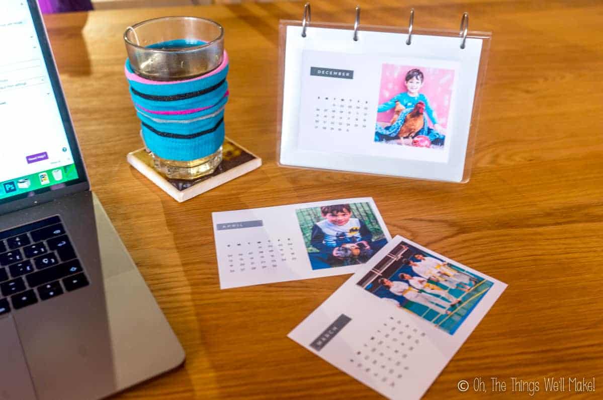 A homemade photo calendar next to a computer and a glass full of tea covered in a blue sock cozy.