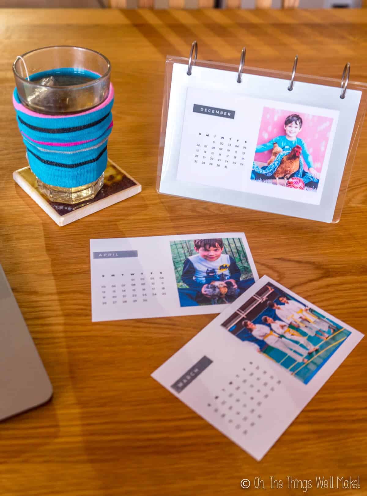 A homemade photo calendar in a flip frame next to a computer and a glass full of tea and some extra printed out photo calendars.