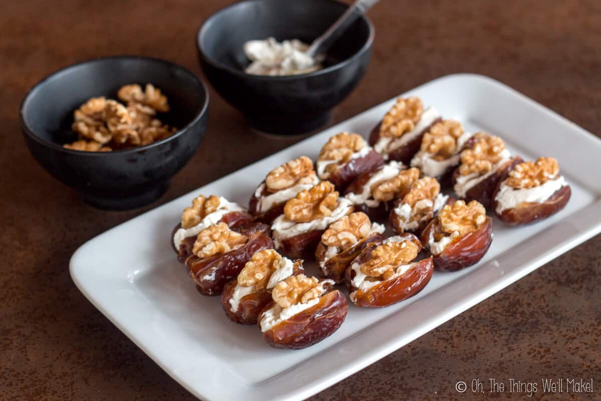 A rectangular plate with sixteen dates laid out. Dates are cut open in the middl, and filled with cream cheese and topped with walnuts. Behind the plate are two bowls, one filled with halved walnuts and another with cream cheese.