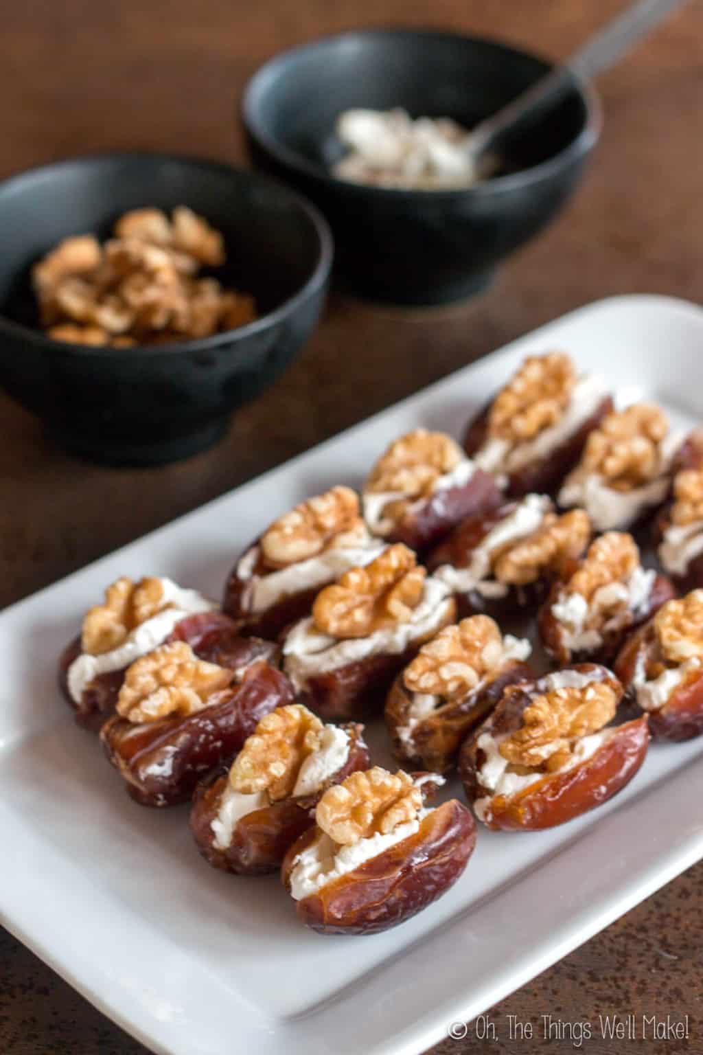 A rectangular plate with sixteen dates laid out. Dates are cut open in the middl, and filled with cream cheese and topped with walnuts. Behind the plate are two bowls, one filled with halved walnuts and another with cream cheese.