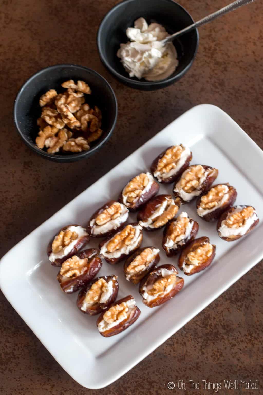 Overhead view of a rectangular plate with sixteen dates laid out. Dates are cut open in the middl, and filled with cream cheese and topped with walnuts. Behind the plate are two bowls, one filled with halved walnuts and another with cream cheese.