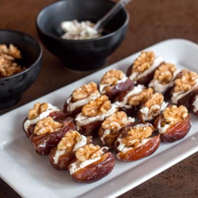 A rectangular plate with sixteen dates laid out. Dates are cut open in the middl, and filled with cream cheese and topped with walnuts. Behind the plate are two bowls, one filled with halved walnuts and another with cream cheese.