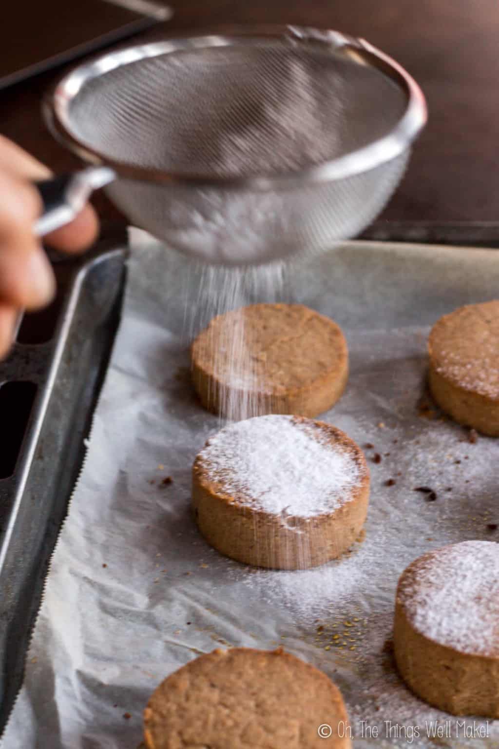 Close up of a metal strainer filled with powdered sugar being sprinkled on freshly baked mantecados and polvorones.
