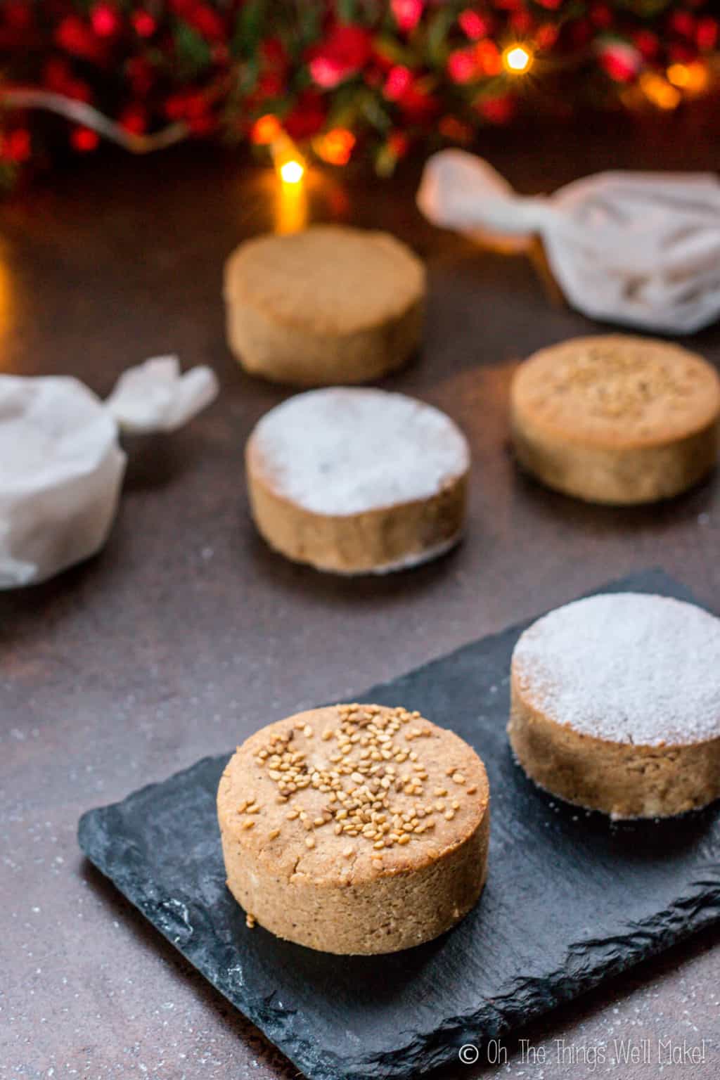 A close up of one mantecados and one polvorones cookies on a black slate platter, with other cookies on the countertop (some cookies wrapped in parchment paper.)