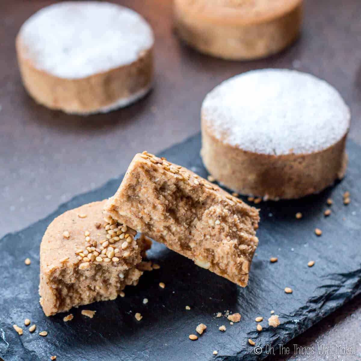 Close up of a mantecados broken in half beside a polvorone topped with powdered sugar, on a black slate platter.