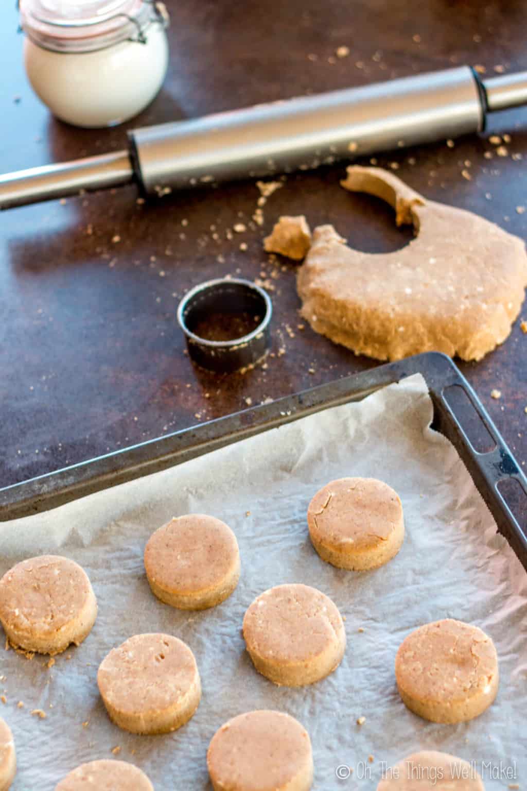 Several pieces of cut mantecados on a baking sheet. On top is a rolling pin, dough with a cut section and a circle cutter.