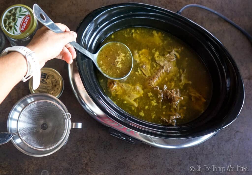 Overhead view of a turkey broth in a slow cooker being spooned up into a ladle, ready to be strained into a jar.