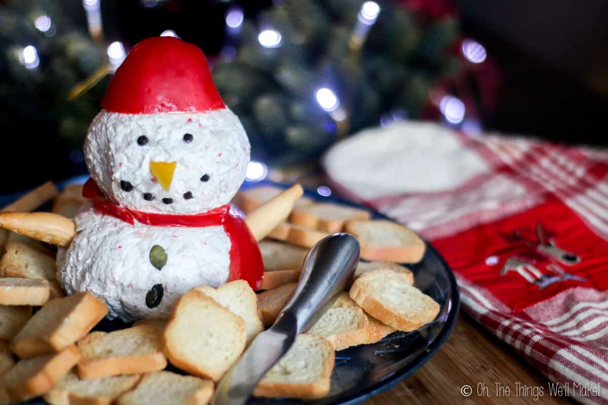 A homemade snowman cheese ball on a platter surrounded by crackers next to a Christmas towel.