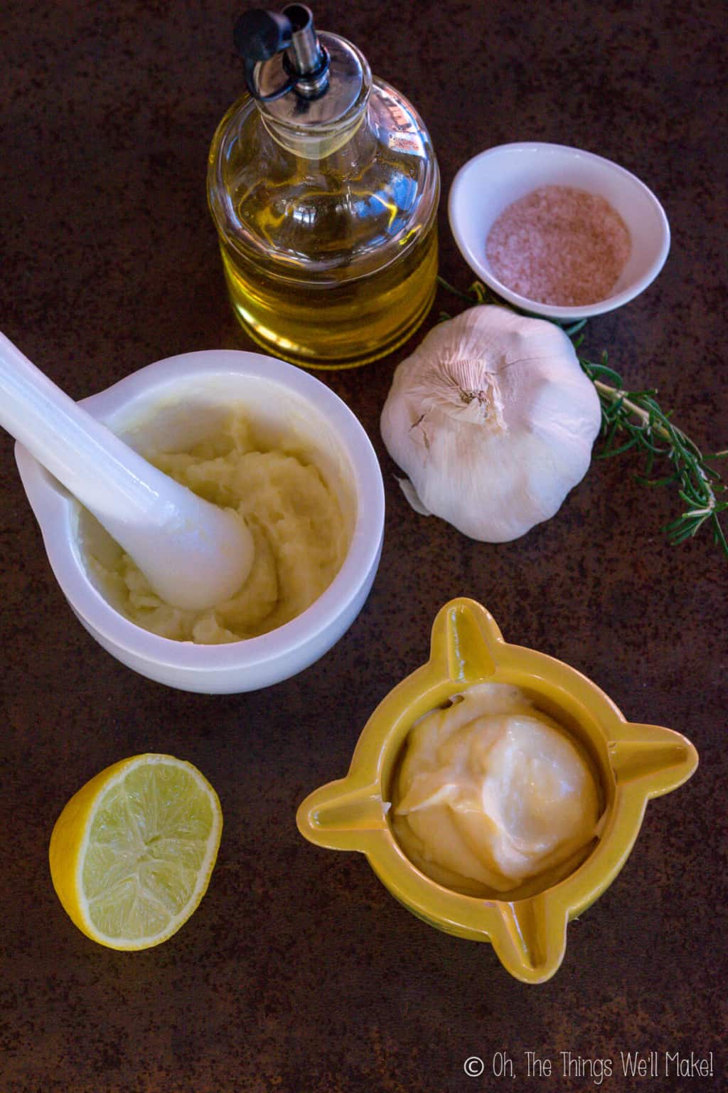 Traditional aioli in a white mortar next to a garlic head, bottle of oil, and a small bowl of pink salt. At the bottom is modern aioli in a yellow mortar with a sliced lemon on its left side.
