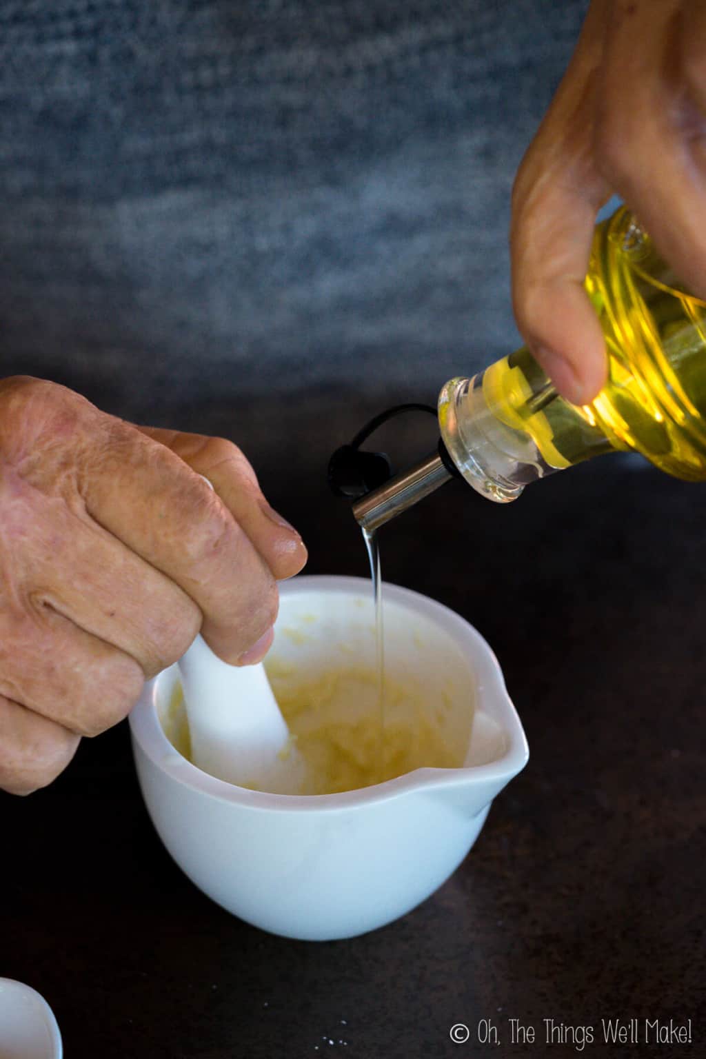 Close up of hands making traditional aioli . One hand holding a pestle grinding aioli, and one hand holding a bottle of olive oil pouring it into the aioli mixture in the white mortar.