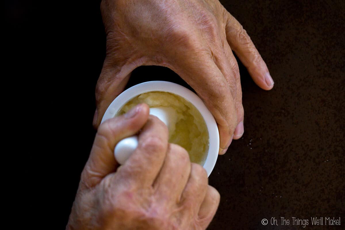 Overhead view of two hands, one holding onto a white mortar and another holding a pestle grinding some aioli.