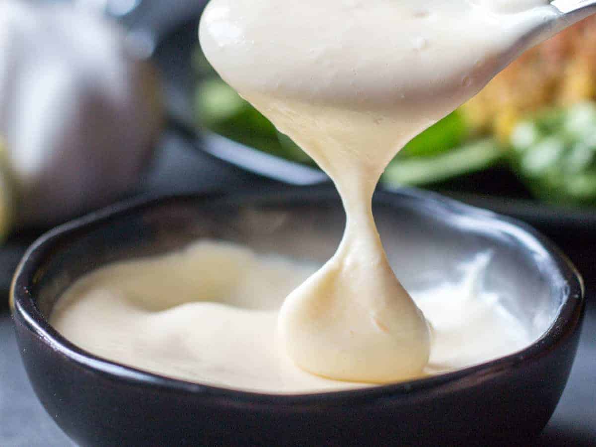 Close-up of a spoonful of homemade white aioli sauce dripping into a black bowl, also full of aioli.