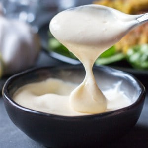 Close-up of a spoonful of homemade white aioli sauce dripping into a black bowl, also full of aioli.