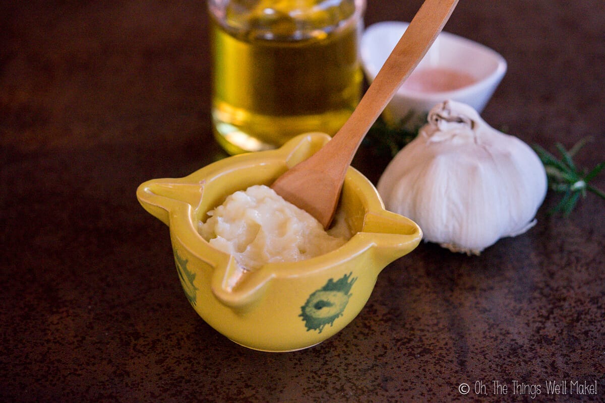 Traditional aioli inside a traditional yellow Spanish mortar with a whole garlic head beside it, and oil and salt behind it.
