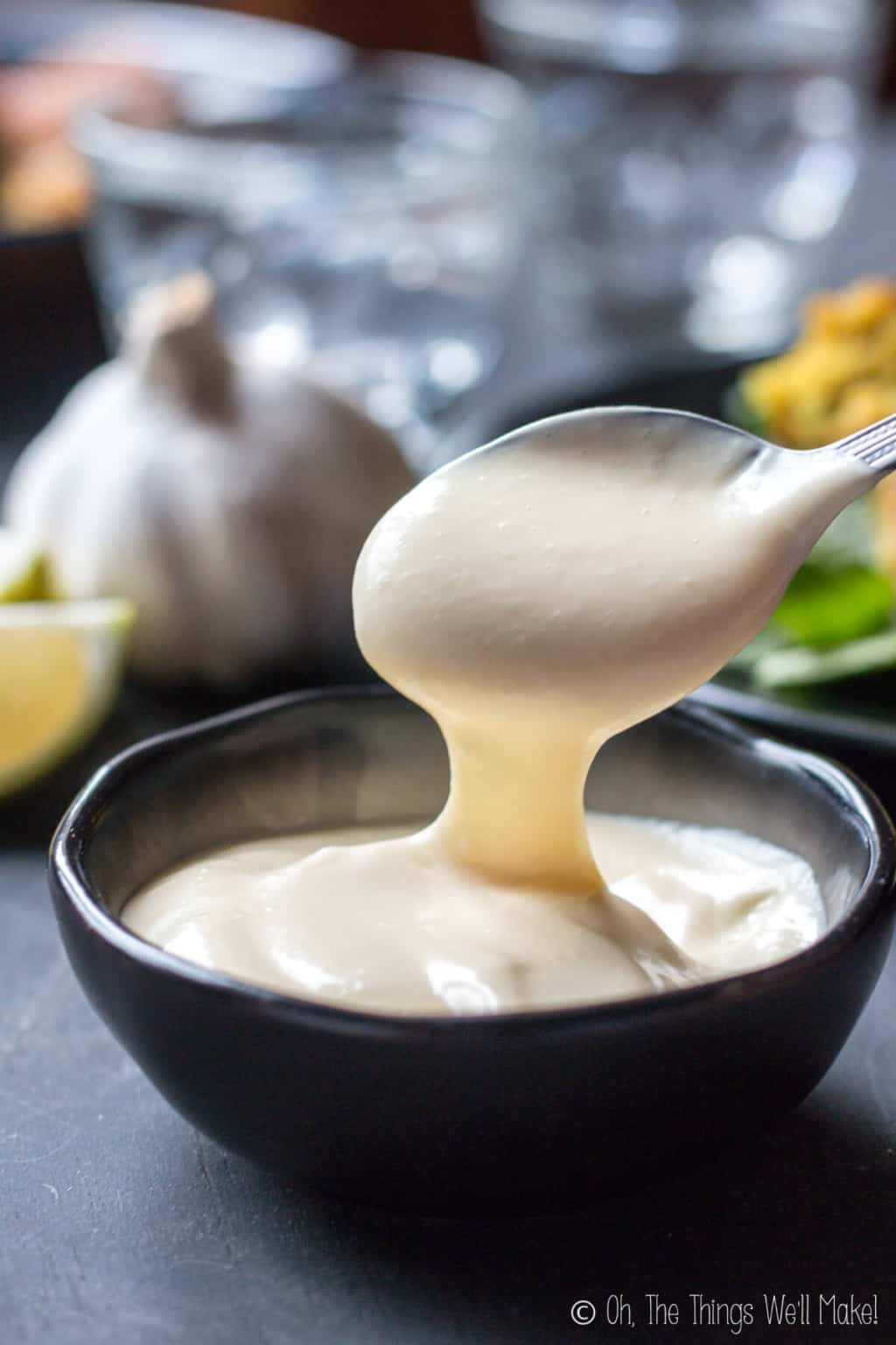 Close up of a spoon full of homemade white creamy aioli lifted on top of a black bowl filled with the same aioli.