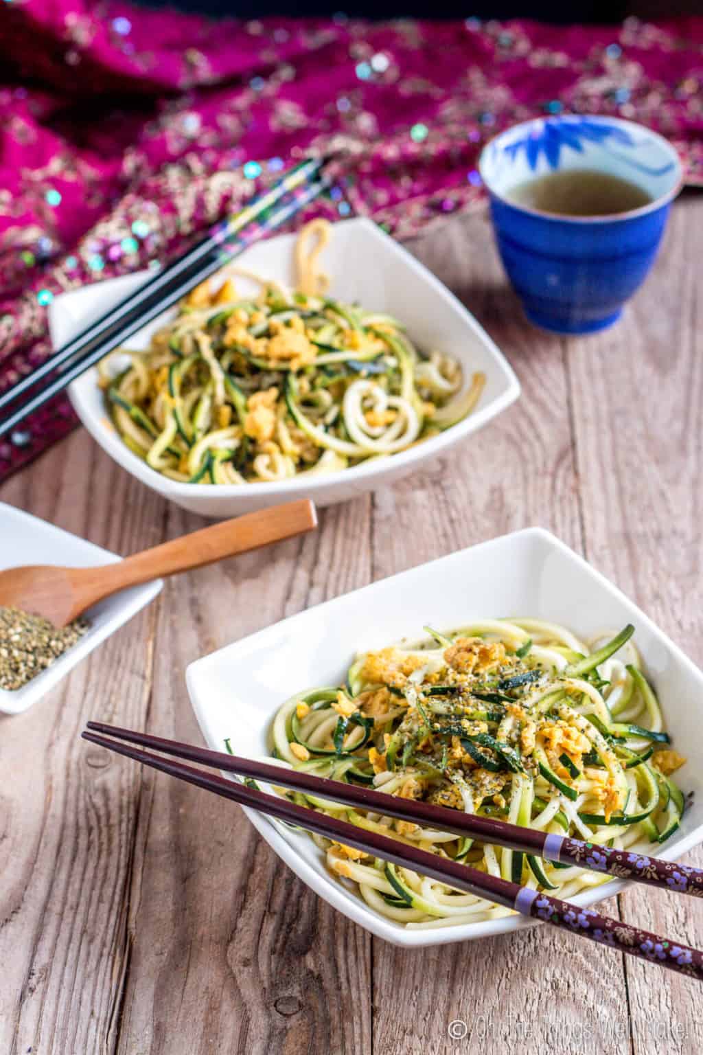 Two white square bowls filled with  pan fried zucchini noodles garnished with eggs, and have black chopsticks on the side of the bowls.