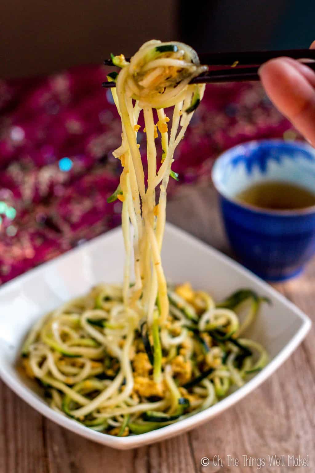 Close up of zucchini noodles being held over a bowl with a pair of black chopsticks.