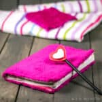 A pink fuzzy covered notebook and a pencil with foam hearts placed on a wood pallet, with pink striped cloth scraps in the background.