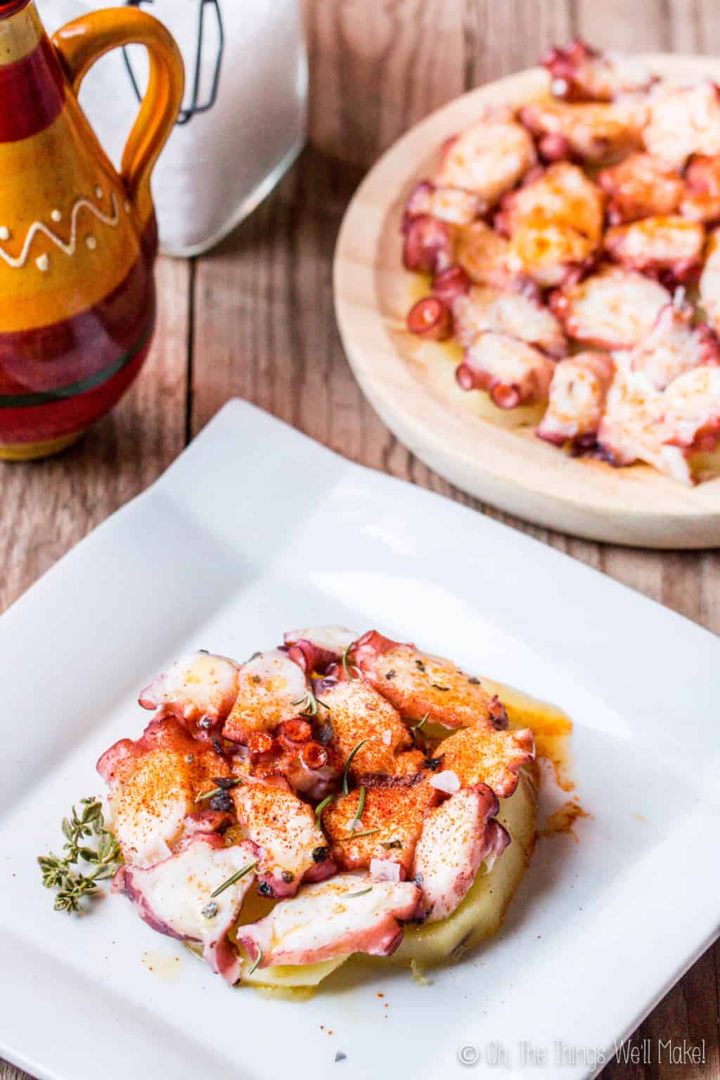 A white square plate with pulpo a la gallega, galician style octopus on top of slices of potato, garnished with paprika on it. On top is a circle plate full of pulpo a la gallega.