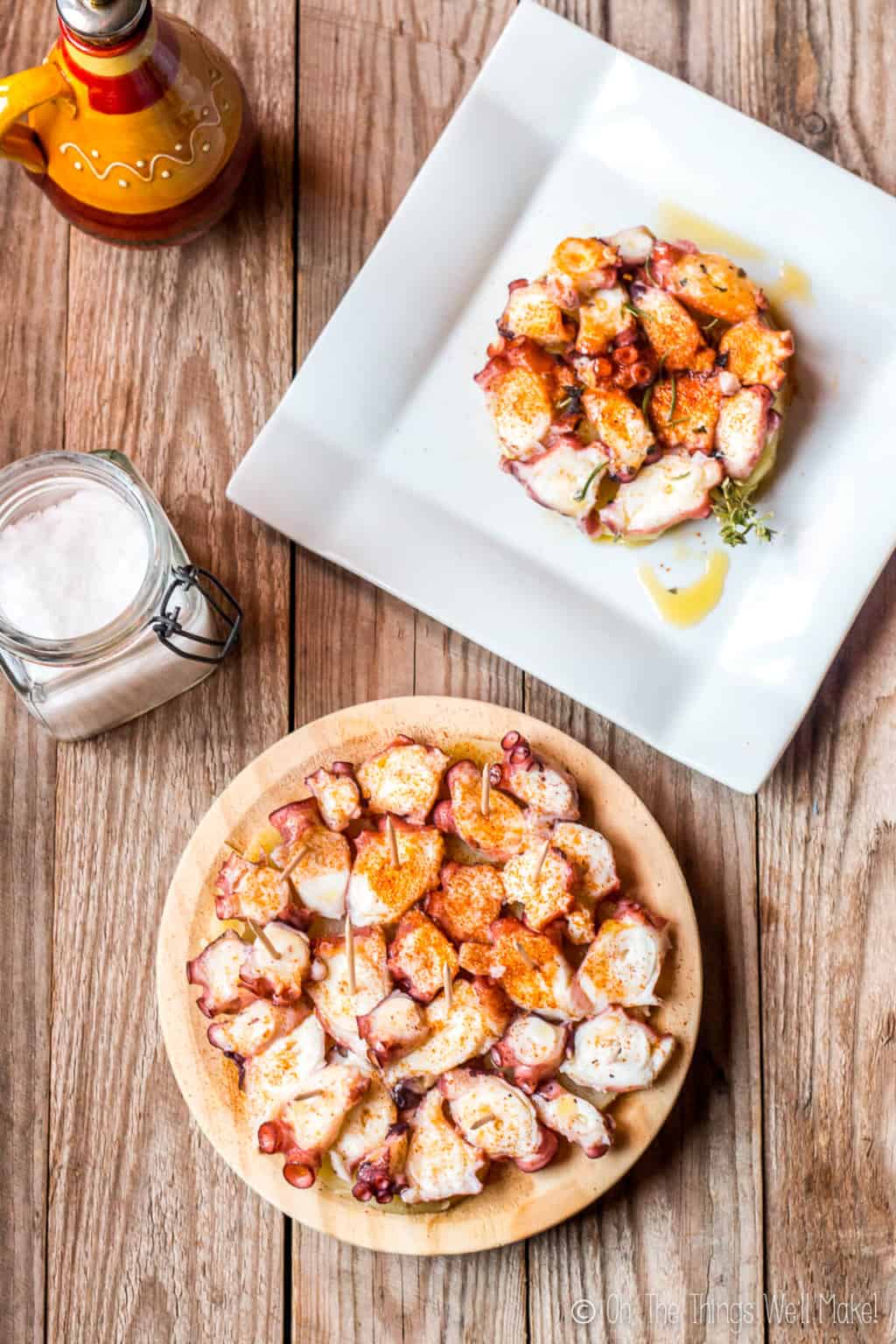 A white square plate with pulpo a la gallega, galician style octopus on top of slices of potato, garnished with paprika on the top. On the bottom is a circle plate full of pulpo a la gallega.