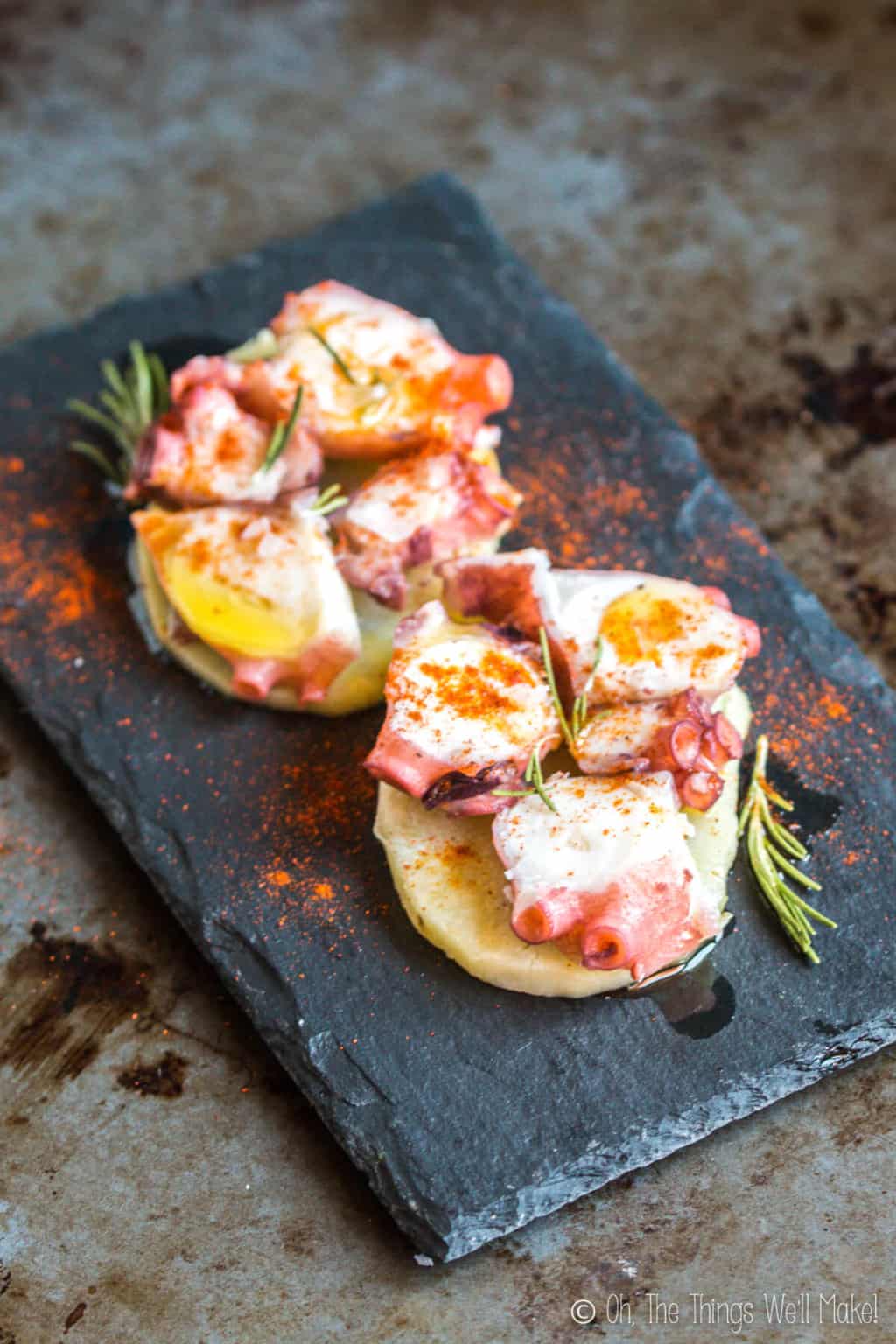 Close up top view of two servings of pulpo a la gallega, galician style octopus on a single slice of potato, placed on a black slate platter.