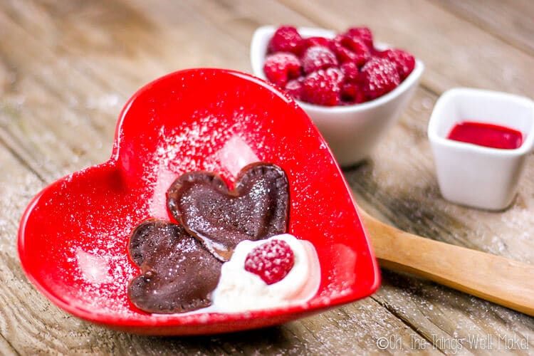 Two pieces of heart shaped chocolate ravioli served in a red heart shaped bowl with whipped cream and fresh raspberries. Beside this bowl is a white small bowl full of raspberries.