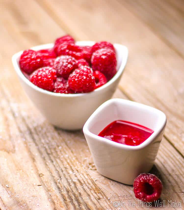 Two white bowls, large bowl filled with red raspberries and small bowl filled with homemade raspberry puree, placed on a wood surface.