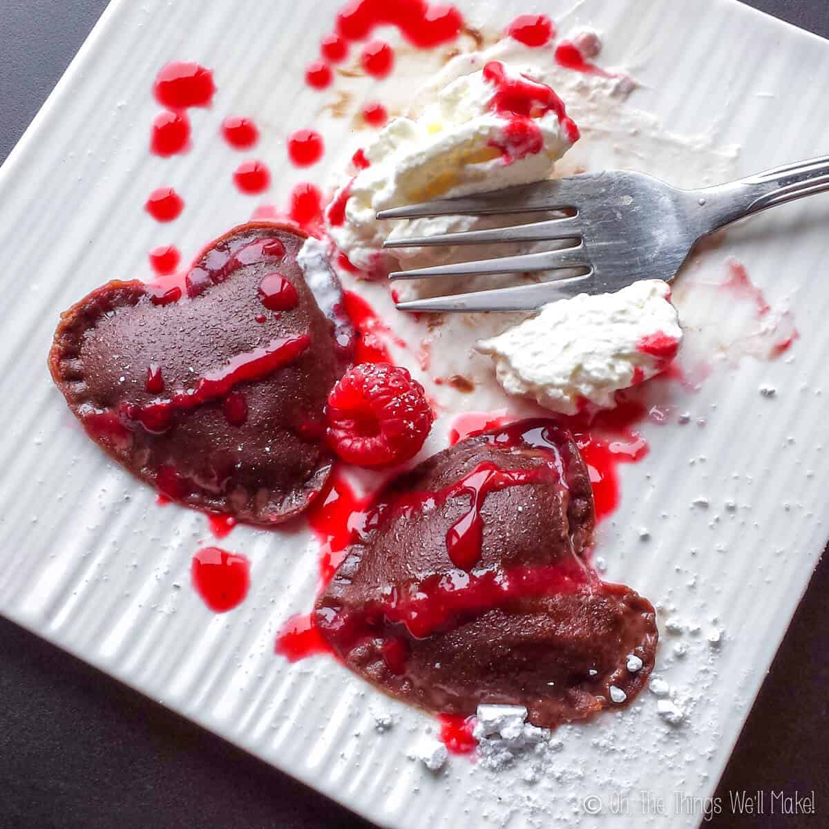 Top view of partly eaten plate of heart shaped chocolate ravioli served with whipped cream, raspberries and raspberry sauce.