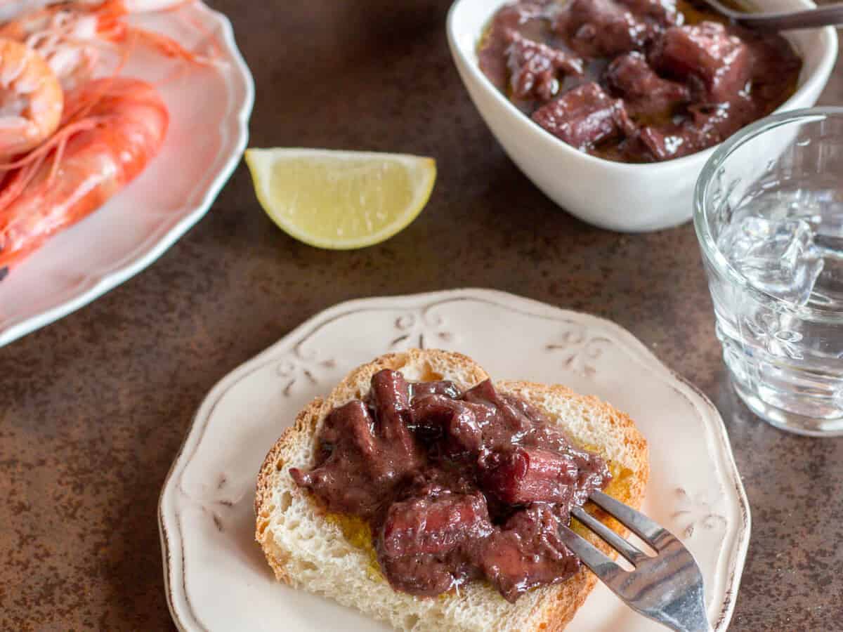 A plate of red wine braised squid on top of a slice of bread with a plate of shrimp, slice of lemon, and a bowl of squid in the background.