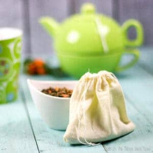 Closeup of a homemade cloth teabag with a small bowl behind it filled with a loose tea. In the background, there is a green teapot and a teacup.