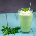 A glassful of homemade green shamrock shake topped with whipped cream and a clover leaf, with a stainless steel straw inside. A bunch of clovers laid down next to the glass. All placed on top of a blue wood tabletop.