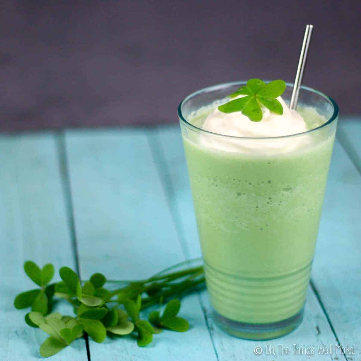 A glassful of homemade green shamrock shake topped with whipped cream and a clover leaf, with a stainless steel straw inside. A bunch of clovers laid down next to the glass. All placed on top of a blue wood tabletop.