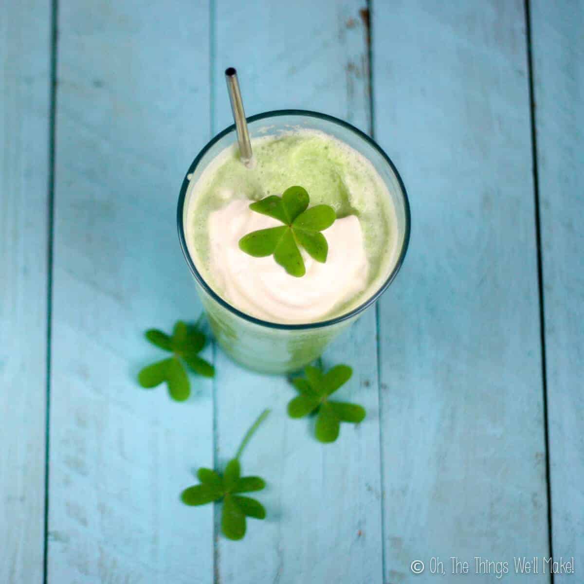 Overhead view of homemade shamrock shake topped with whipped cream, clover leaf, and served with a stainless steel straw. The clover leafs are on the table beside it.