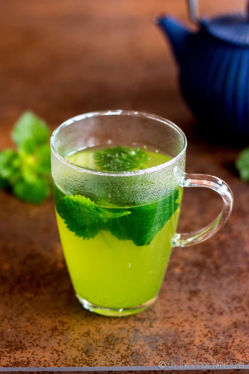 Freshly brewed green stinging nettle herbal tea in a glass in front of a blue cast iron teapot.